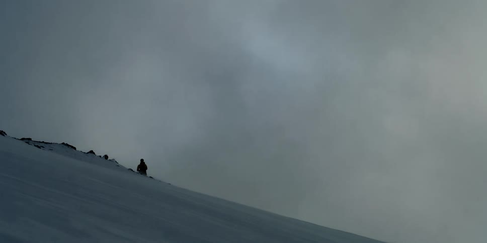 A man stands on a snowy slope as clouds fly by over the sun