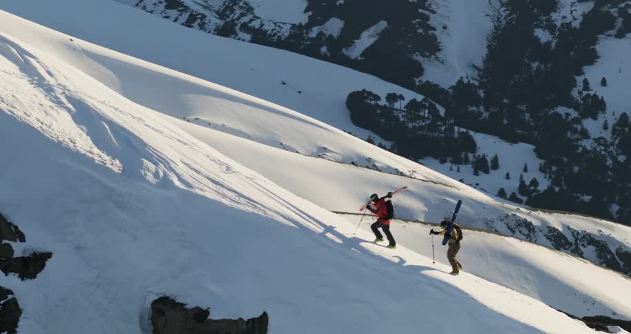 Aerial of two people climb a mountain ridge with skis