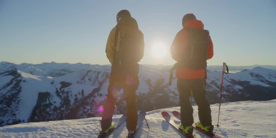 Two skiers stand at the edge of a snowy drop off at sunrise