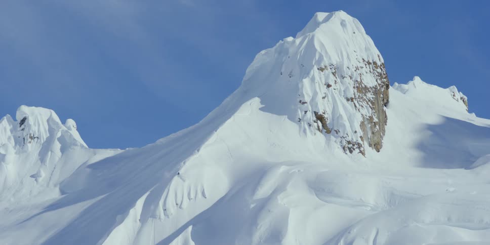 A skier skis down the ridge of a steep mountain slope on a bright blue sky day