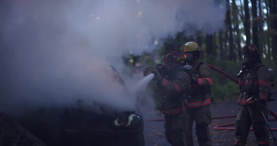 A group of firefighters using a fire hose to spray the smoking wreckage of a car