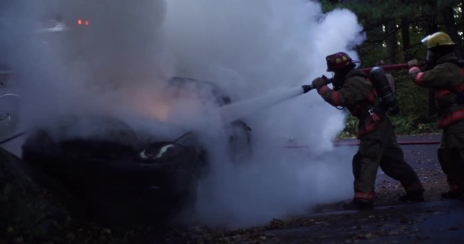 Fireman hosing down a car engulfed in flames in a wooded area