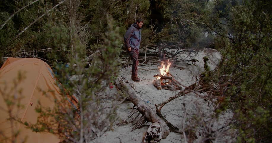 Man stands in front of a campfire near a tent in the forest