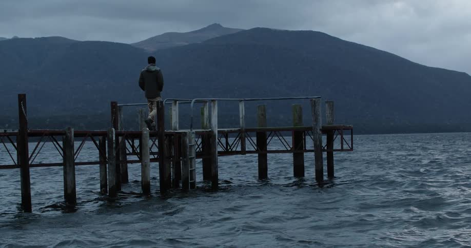 A man walks toward the edge of a wooden dock surrounded by a lake and mountains