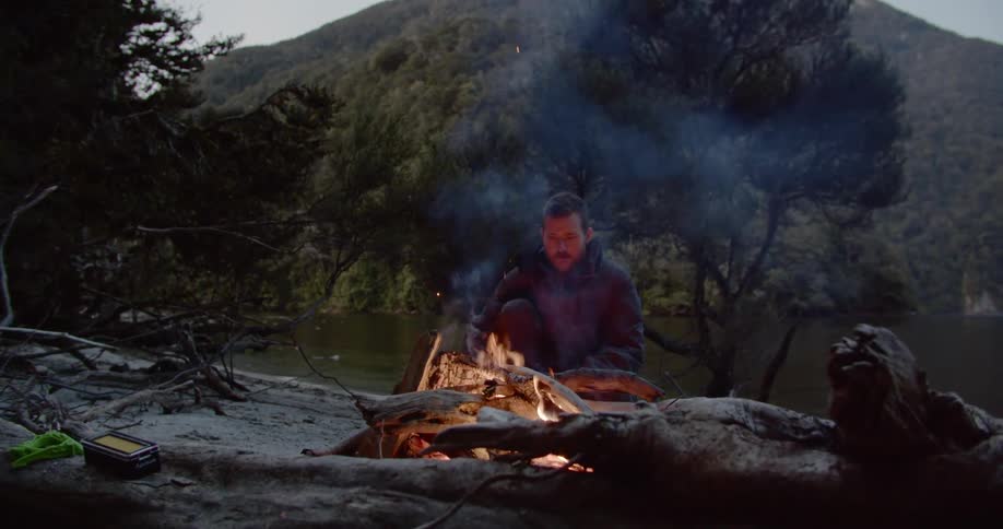 Man sits in front of a campfire on the shore of a lake in the mountains
