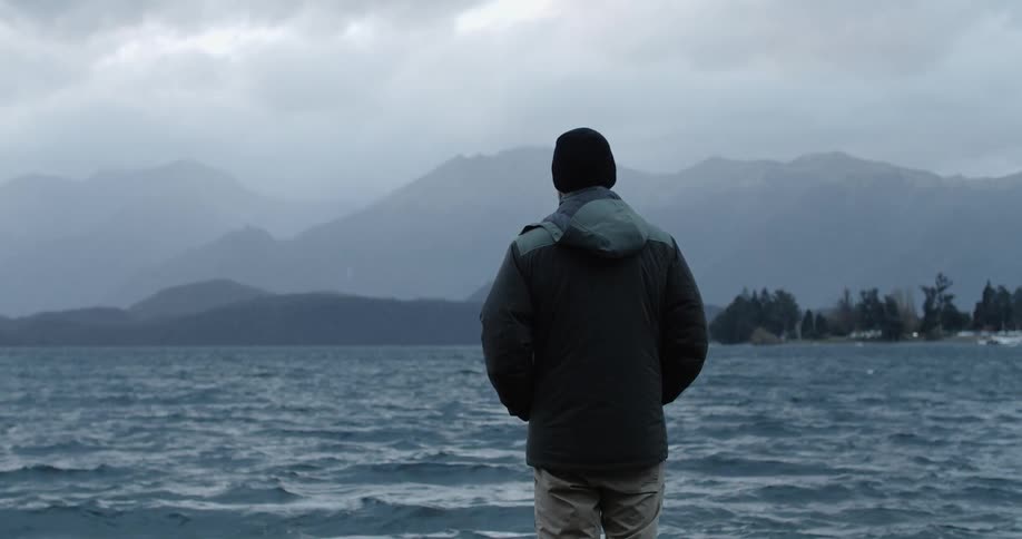 A man in a coat and a hat stands by the shore of a lake