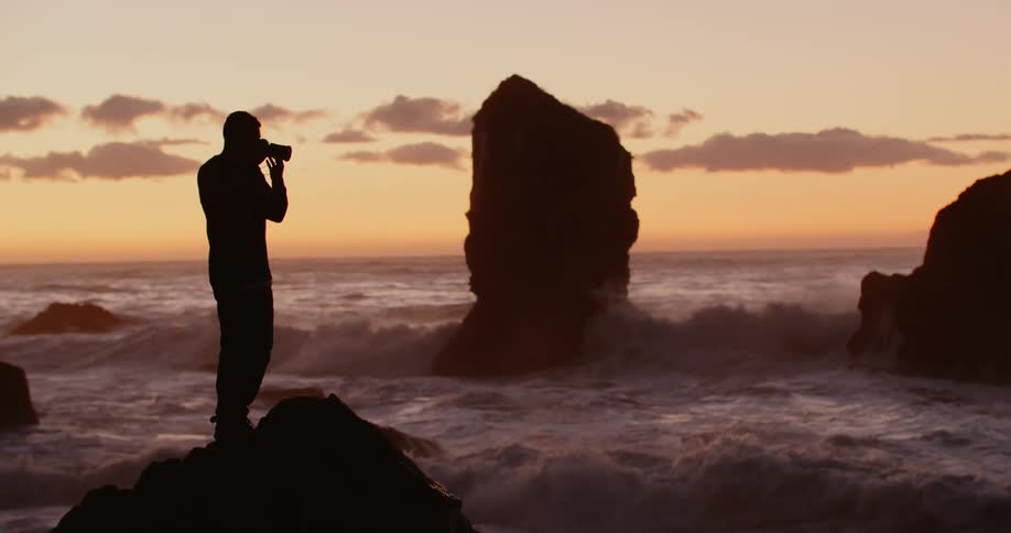 A man with a camera stands on a rock at the beach and takes a picture of the ocean