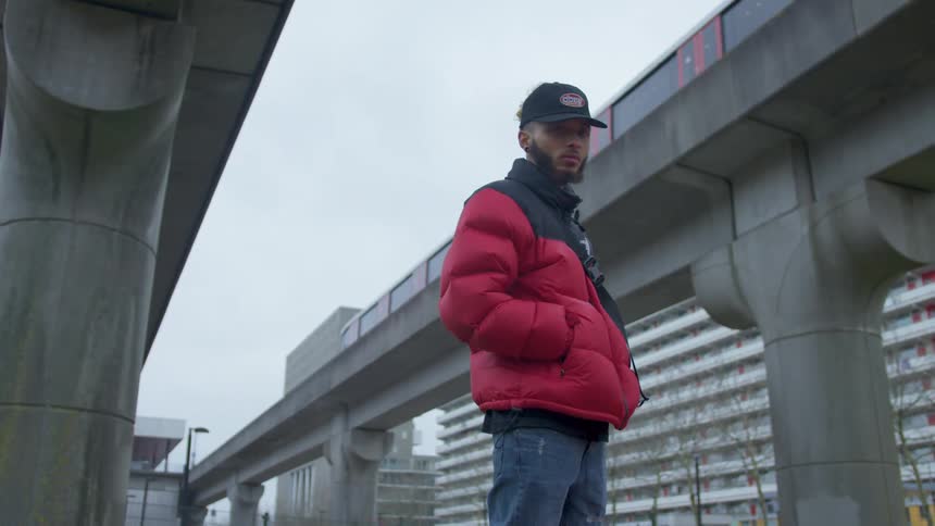 A black man in a coat and a hat stands outside by the train overpass in a city
