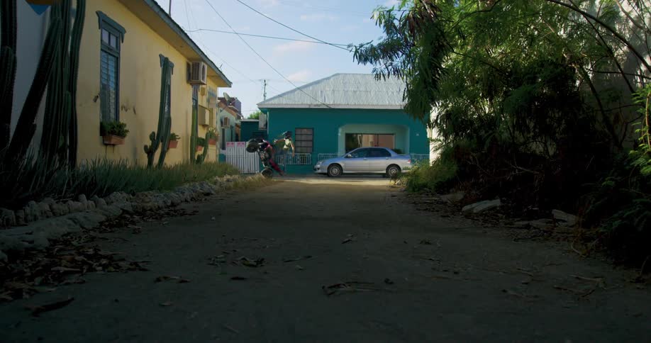A person rides a bike down a dirt alley by small home