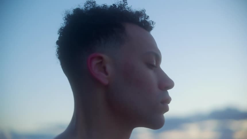 A bare chested man stands outside in profile shivering
