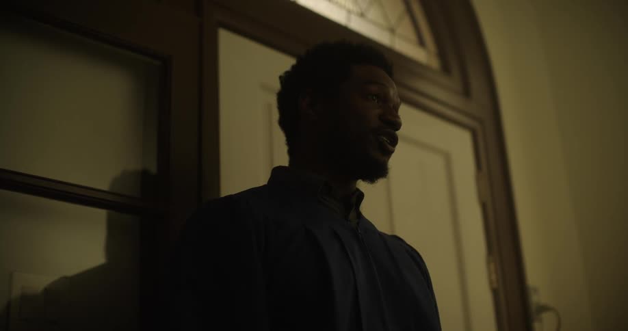 A black man in a robe stands inside a room and talks