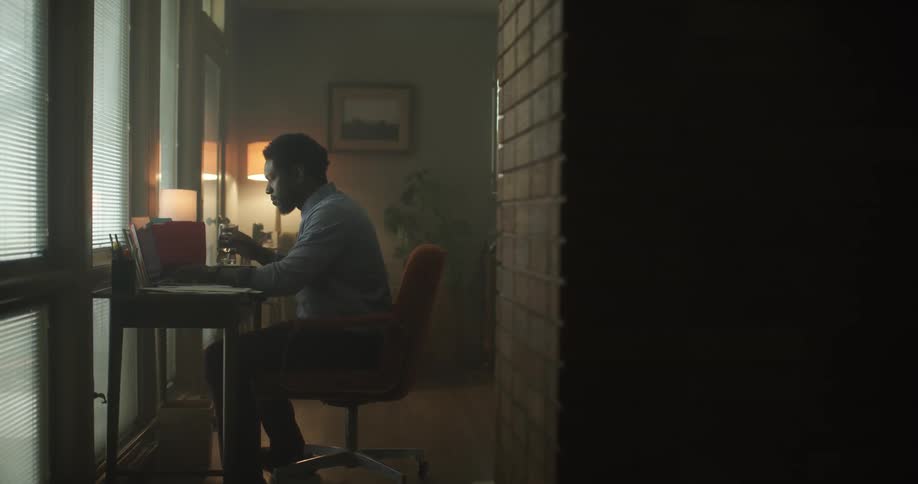 A black man sits at a desk with a drink and a laptop