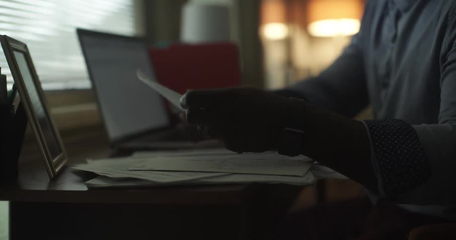 A man is typing on his laptop and pauses to read a piece of paper on his desk