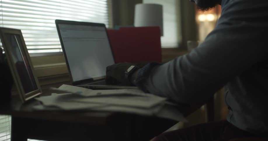 A man sits at a desk in his home typing on a laptop