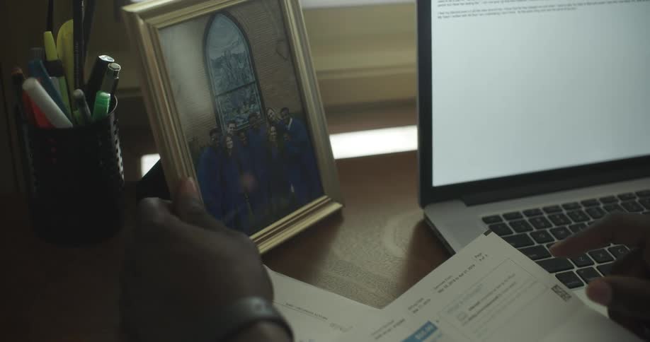 A person sits at a desk in front of a laptop and reaches out to touch a picture frame