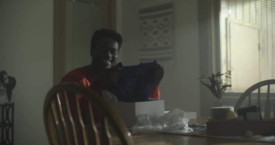 A black man sits at his kitchen table and smiles at a robe in an opened box