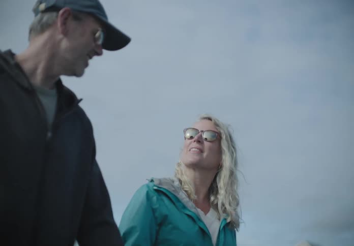 An older white couple hold hands and walk outdoors on the beach