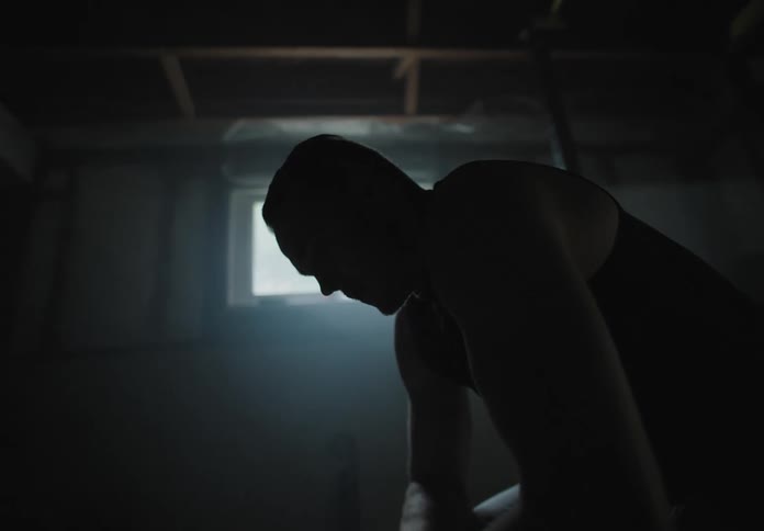 A large muscular man sits and then gets up and walks out of the basement