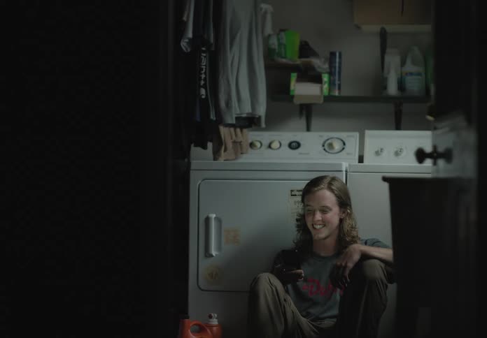 A white man sits on the floor of a laundry room talking on a smartphone