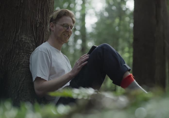 Male sitting against a tree trunk in the woods while resting a smart phone on his bended leg and video chatting