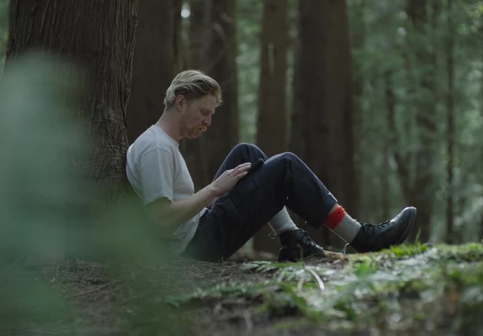 A man sitting on the ground leaning on a tree trunk tapping on his phone
