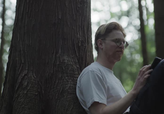 A white man with glasses and a mustache leans against a tree and speaks while touching a smartphone