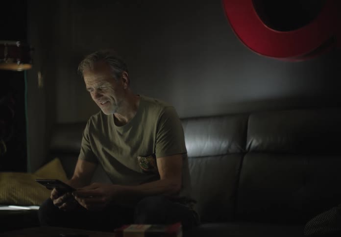 An older white man sits on a couch fiddling with a smartphone