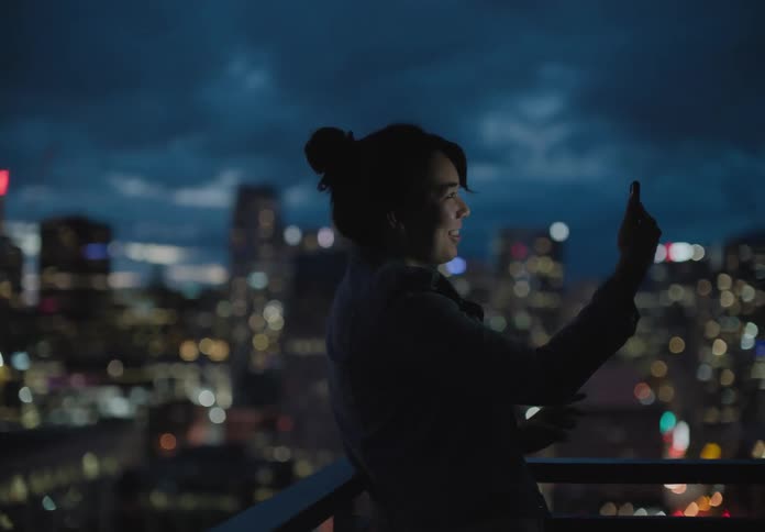 Female standing outside leaning against the balcony as she holds her cell while video chatting with the city lit up behind her under a dark cloudy sky