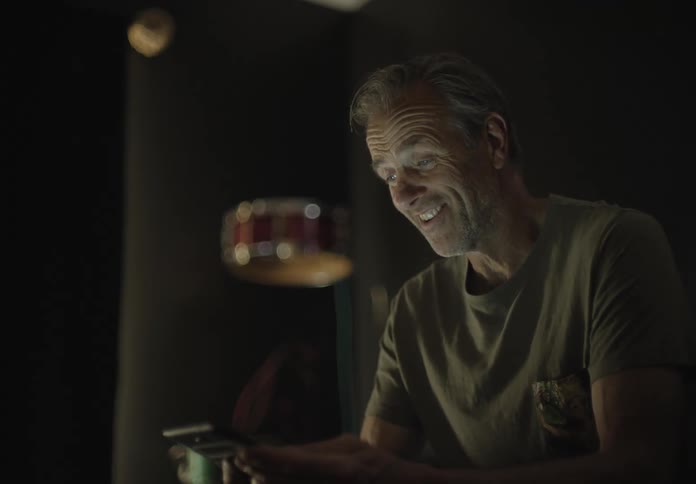 A white man sits in a room in the dark while talking to someone through his smartphone