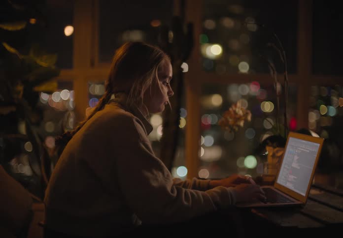 A woman sits down at a desk and laptop next to windows showing a dark city
