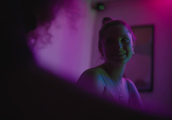 A white female teenager smiles while talking animatedly to another person in an apartment