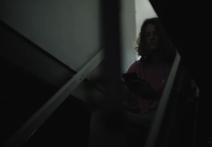 Male walks through a basement doorway and down three steps before he sits down and starts video chatting on his cell phone