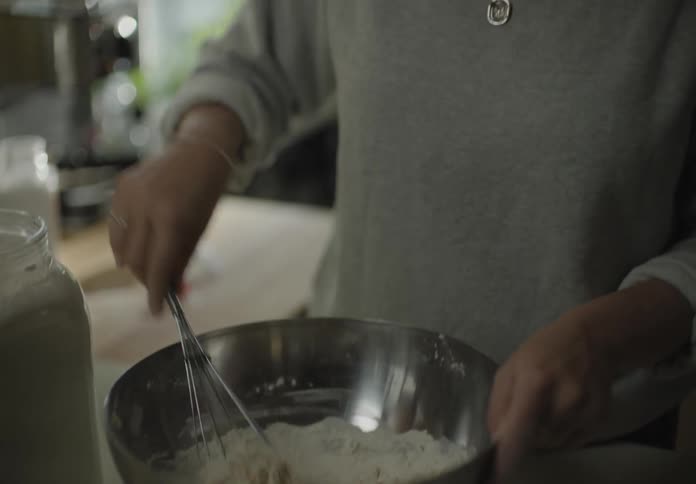 Female standing in front of kitchen counter mixing together ingredients in a steel bowl with a whisk and she talks and smiles