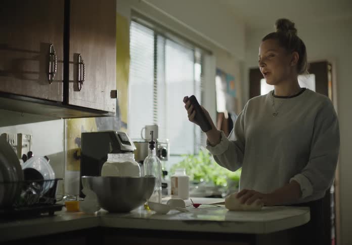 Young woman kneading dough in kitchen takes video cal on her smart phone