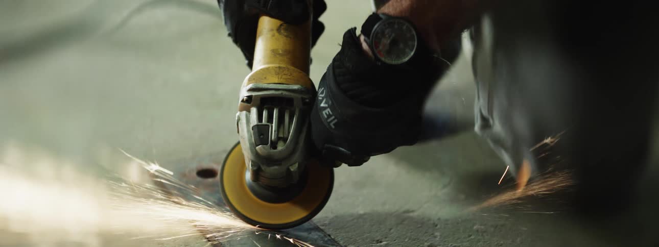 A kneeling person using an angle grinder on a piece of metal on the ground causing sparks to fly