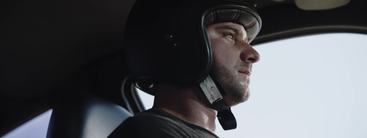 A man wearing a padded helmet drives a sports car on a sunny day