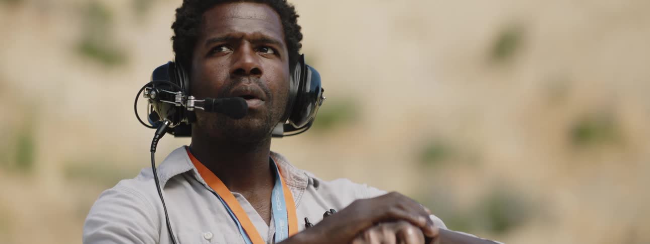 A man wearing a radio headset stands with his hand on his watch to time a car racing by