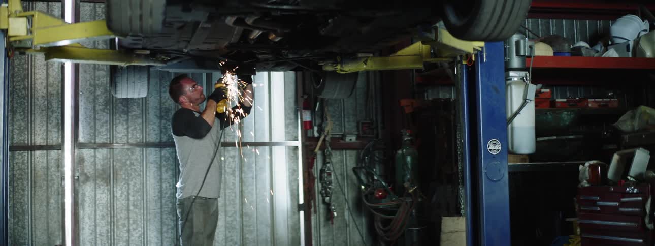A man using a power tool on the bottom of a car lifted by a car lift in a garage