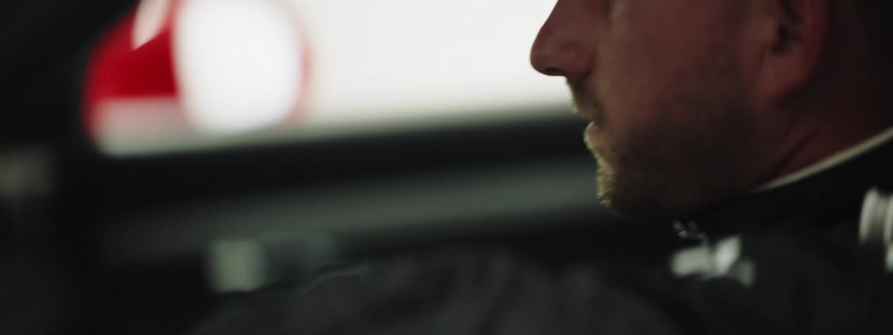 Male race car driver picks up a red helmet sitting in the drivers seat and puts it on his head to prepare to race the car he is sitting in