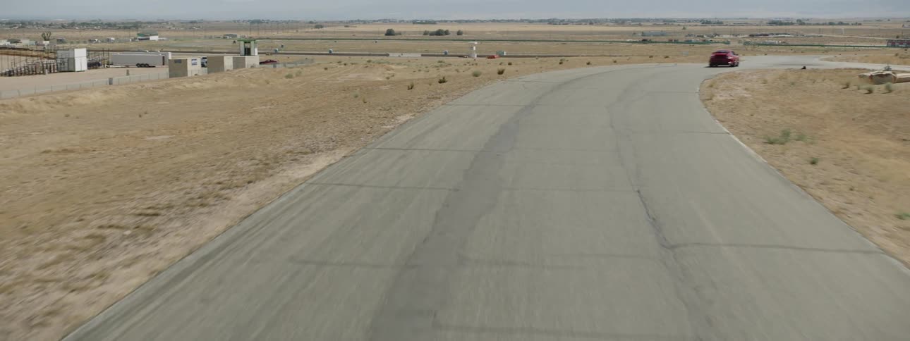 Red sports car races down winding road between fields near buildings