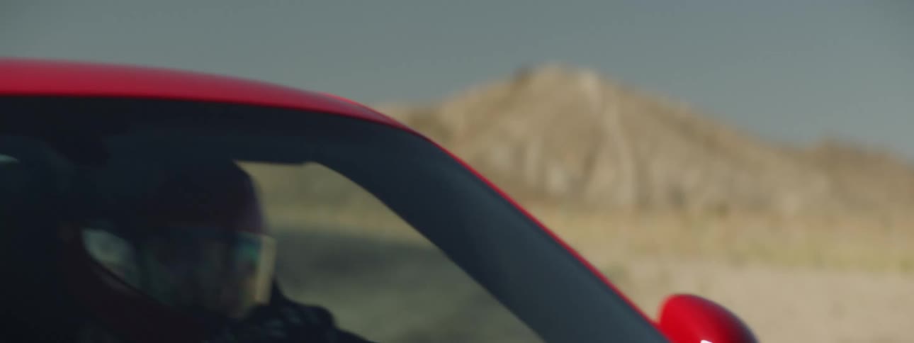 A driver in helmet and visor drives a red sports car on an empty desert track
