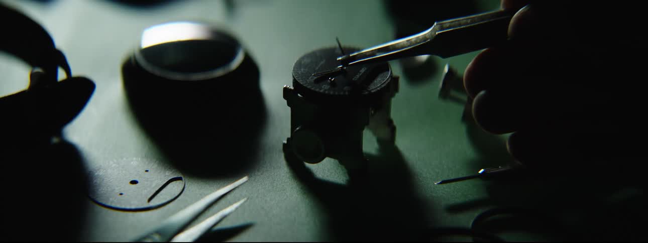 A hand with tweezers removes one of the hands from a watch on a table with parts and tools spread out