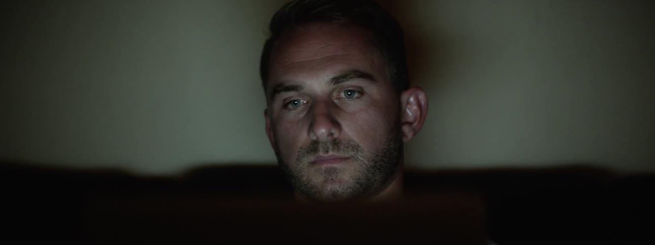 A white man sits in a room and watches television at night