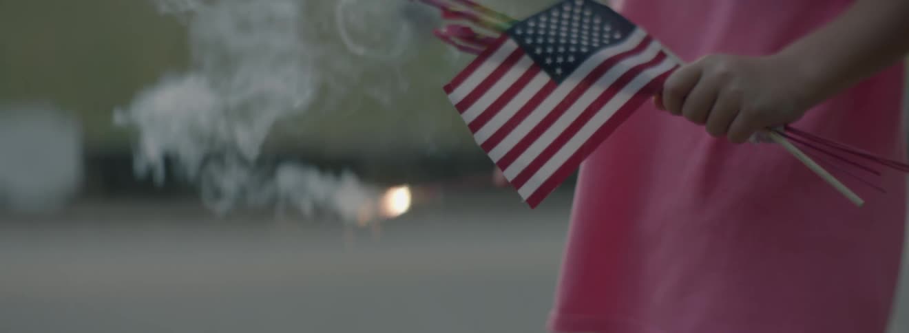 A girl holds a burning sparkler and american flag outside in the summer