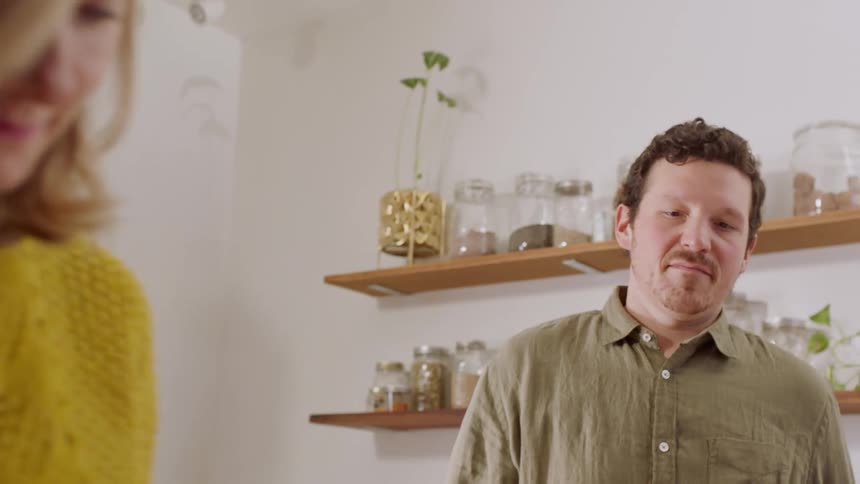 A man watches as his wife prepares plates of food in their kitchen