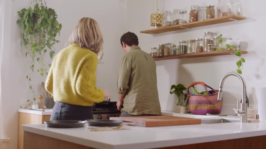 A woman sets plates on the counter as a man cooks at the stove in a bright kitchen