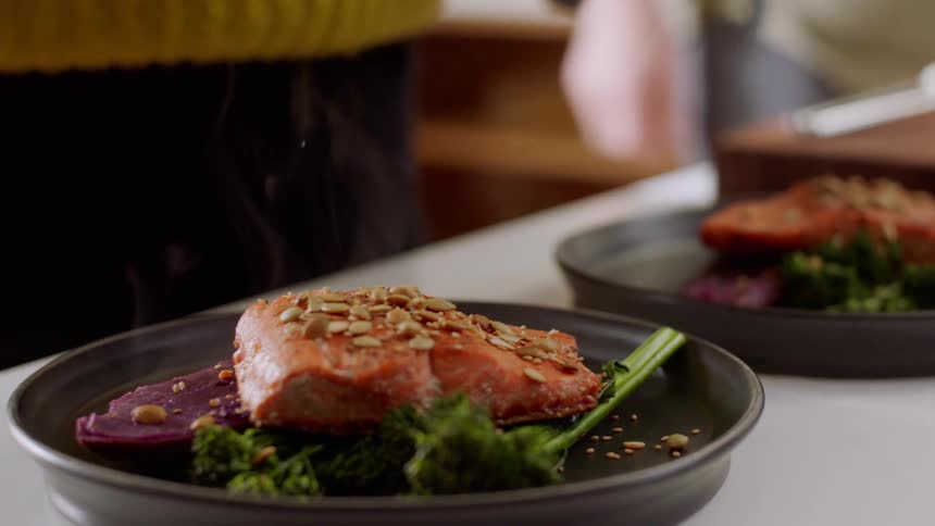 A white woman sprinkles seasoning on plates of salmon and vegetables