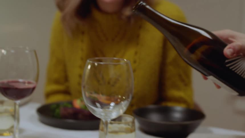 Man pours two glasses of wine at dinner table in front of young woman