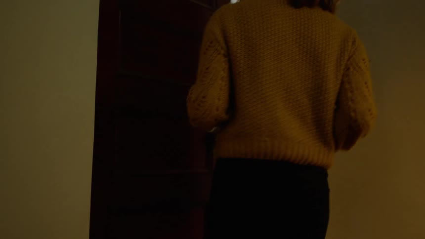A woman walks down the stairs in her house holding a pot of coffee and goes into the living room