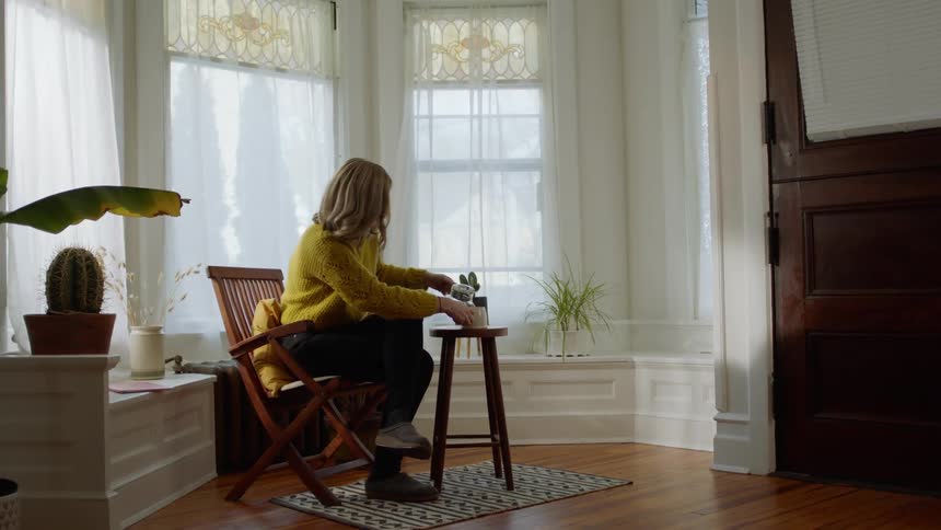 A white woman walks over to a chair and sits down and pours herself a drink